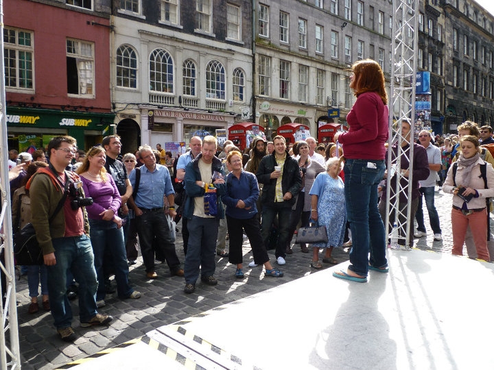 Hannah stood on a stage on cobbled stones performing to a large gathered crowd during the Edinburgh Fringe Festival